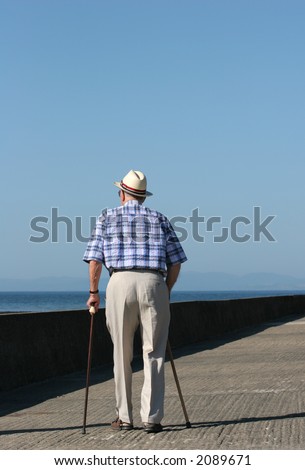 Rear view of an elderly disabled man walking with two walking sticks on a beach promenade.