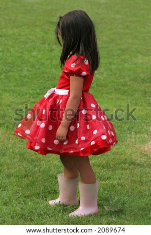 Little girl in a red and white polka dot dress wearing pink wellington boots and standing sideways on the grass.