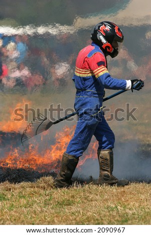 Man in a multi colored boiler suit, wearing a crash helmet, carrying a rake and trying to put out a fire on the grass.