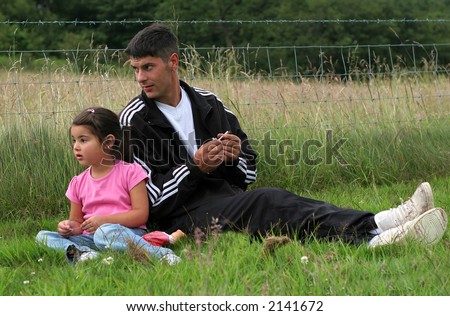 Man and a little girl sitting together on the grass looking at something in the distance with a wire fence and field to the rear. Man is holding a cigarette in his hand.