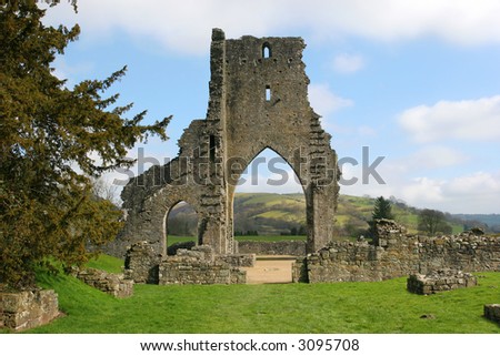 Talley Abbey monastery ruins with rural countryside beyond the main arch and grass in the foreground. Set against a pale blue sky with clouds.