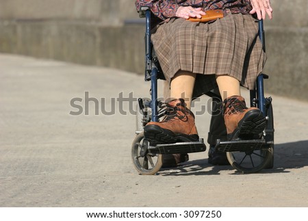 Lower body of a an elderly woman sitting in a wheelchair on a pavement, with arthritic hands and wearing large boots.