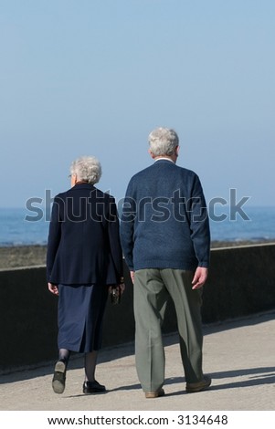 Elderly couple walikng together on a seaside promenade with a blue sky and sea, out of focus, to the rear.