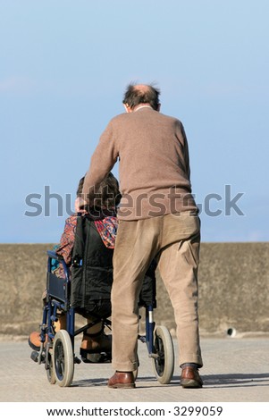 Rear view of an elderly man pushing an elderly female in a wheelchair on a pavement with a blue sky to the rear.