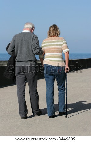 Elderly man helping a middle aged female to walk with a walking stick, with a blue sky and the sea , out of focus, to the rear.