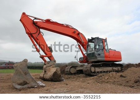Large orange digger standing idle on muddy earth in rural countryside with another large spare digger bucket in the foreground. Set against a grey cloudy sky.