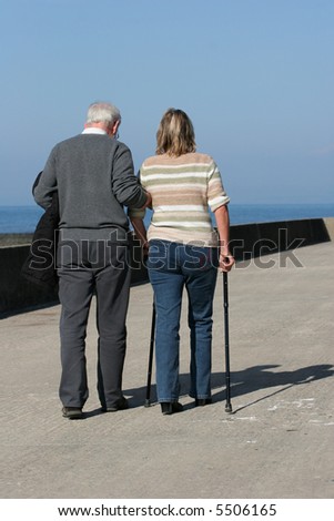 Elderly man helping a middle aged female to walk with walking sticks, with a blue sky to the rear.