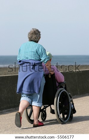Elderly woman pushing a man in a wheelchair on a seaside promenade. Sea (out of focus) to the rear).