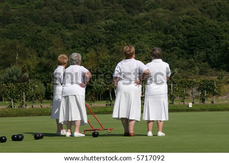 Four elderly females (rear view) dressed  in white lawn bowling outfits and standing on a bowling green. One of the ladies is holding a red metal ball gatherer. Trees and shrubs to the rear.