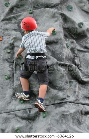 Young boy climbing on a rock training face, wearing a safety harness and red helmet.