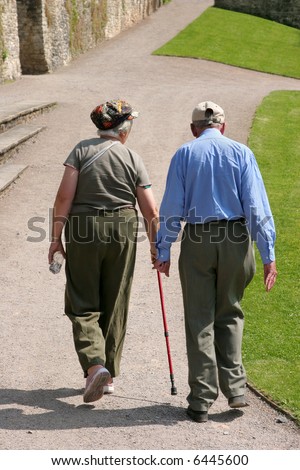 Elderly man and woman walking together along a path with the female holding a walking stick. Rear view.