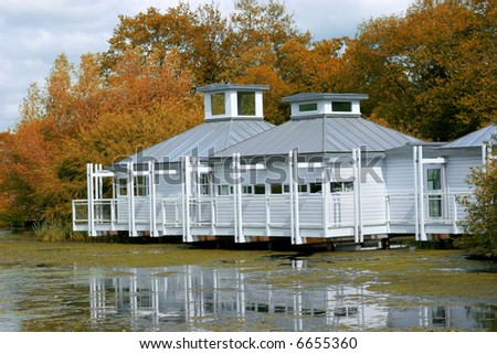 White wooden lakeside residence in autumn with a veranda, with water and pond weed to the foreground and trees to the rear.