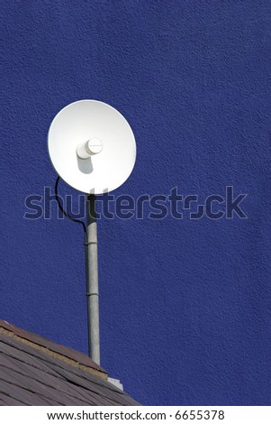 White satellite dish fixed to a roof and in front of a blue pebble dash wall.