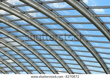 Silver metal curved roof joists in a conservatory with glass paned windows in between and a blue sky with clouds beyond.