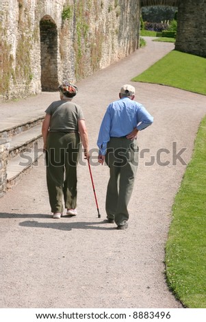 Elderly man and woman walking together along a path with the female holding a walking stick and the man holding his back as if in pain. Rear view.