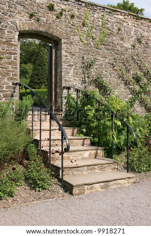 Ancient garden stairway with metal handrails, set within an old stone wall and leading to a garden beyond.  Flowers and shrubs either side of the steps.