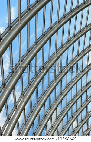 Silver metal curved roof joists in a conservatory with glass panes in between and a blue sky and clouds beyond.