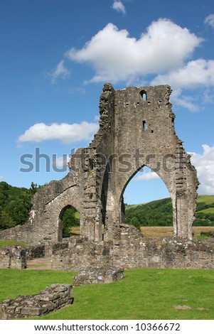 Talley Abbey ruins in Wales, United Kingdom, with rural countryside beyond the main arch and grass to the foreground. Set against a blue sky with clouds.
