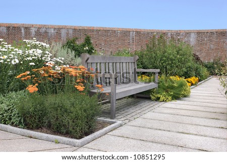 Wooden oak garden bench next to a path, inside a walled garden with flowers and shrubs. Blue sky to the rear.