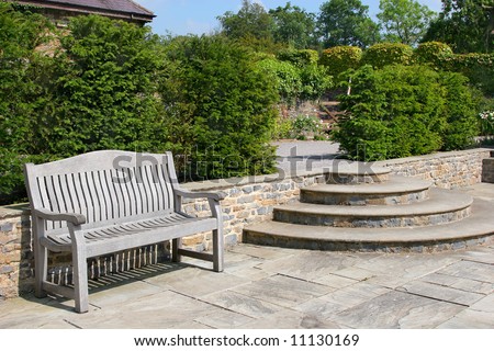 Outdoor garden tiled patio area, with an old wooden oak bench, curved steps to the side and shrubs to the rear.