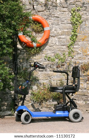 Electric mobility scooter standing idle on a path with an orange lifebuoy hanging on an old stone wall covered with plants, to the rear.