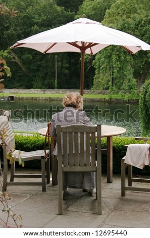 Elderly lady sitting alone at an outdoor wooden table shaded by an umbrella. Lake view and trees to the rear. Rear view of the woman.