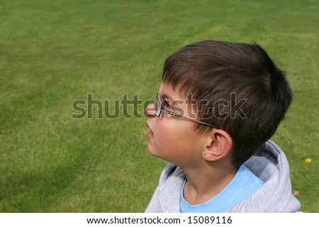 Face of a little boy wearing glasses, with a puzzled look on his face. Grass (out of focus) in the background.