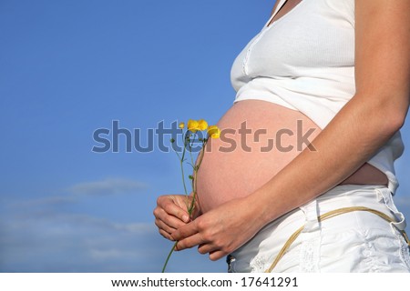 Pregnant woman holding a bunch of buttercups with exposed bare stomach. Blue sky to the rear.