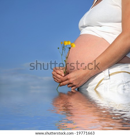 Pregnant woman with exposed bare stomach holding a bunch of buttercups, with reflection over rippled water and a blue sky to the rear.