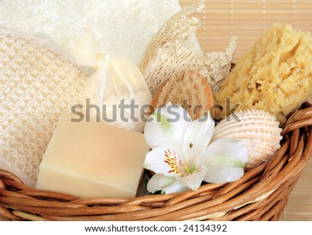 Natural cleansing products in a wicker basket including a cream flannel, soap, sponges, exfoliating sisal pad, hand brush, vanilla bath salts in a net bag and a white lily flower.