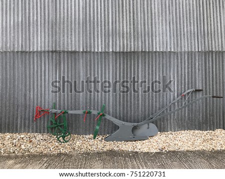 Old metal farming plough on gravel against corrugated iron background.