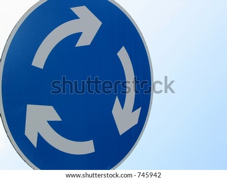 Blue road sign with arrows in a circle against sky. The British roundabout.