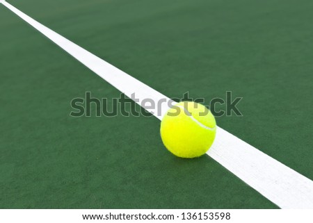 a bright green tennis ball sits on the white line of a green tennis court, diagonal line