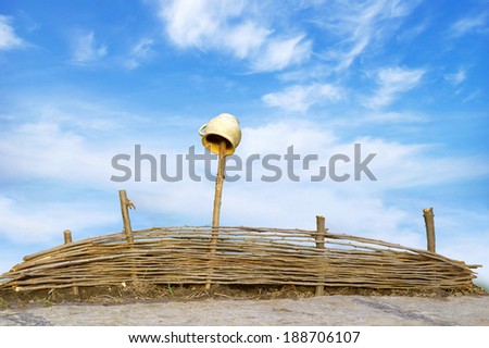 Wicker fence of thin rods with old clay pot hanging to dry on a wooden column, standing near the road, isolated with clipping path on blue sky backdrop with space for text