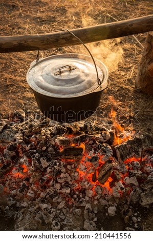 In hanging on a log cast iron cauldron on hot embers fading campfire stew boiling potatoes. Heated smoke rises in warm rays of evening sunset. Close-up view with space for text on blurred background
