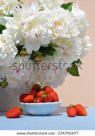 Vivid red juicy sweet strawberries in saucer on the background of a lush bouquet of peonies in delicate white vase with space for text on blue tablecloth
