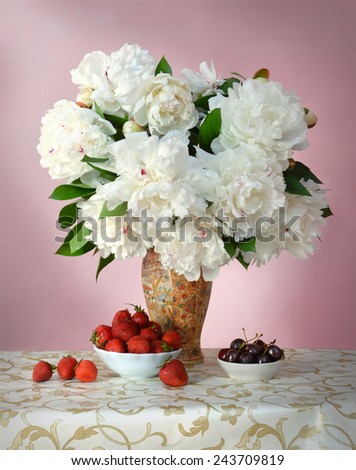 Gorgeous lush white peonies with green leaves in porcelain vase with Chinese painting on openwork tablecloth with golden pattern and dishes with sweet red strawberries and juicy dark burgundy cherries