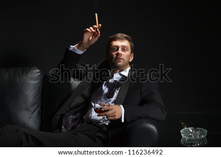 Macho man in a stylish tuxedo sitting in the darkness at a nightclub puffing on a cigar and drinking brandy or cognac