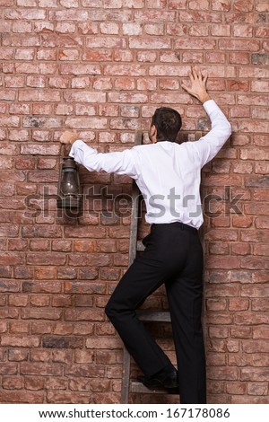 Man at the end of his search up against a brick wall standing balanced at the top of a stepladder with an old oil lamp in his hand with nowhere else to go
