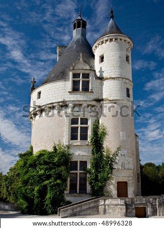A round medieval tower with pointed turrets from Normandy, France.  The tower is white brick and stucco and has green ivy growing on the sides.  Beautiful blue sky with white clouds in the background.