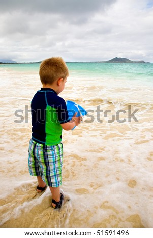 A little boy in a colorful blue and green outfit is playing with a blue bucket at the edge of the water on the beach