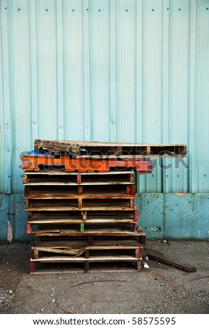A stack of old wooden pallets painted in bright colors, next to the blue corrugated metal wall of an old factory building.