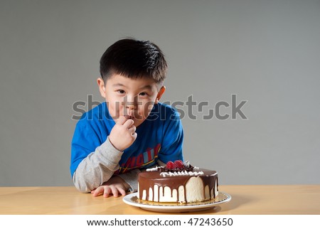 A cute Asian kid posing with a birthday cake, nibbling at the cake.