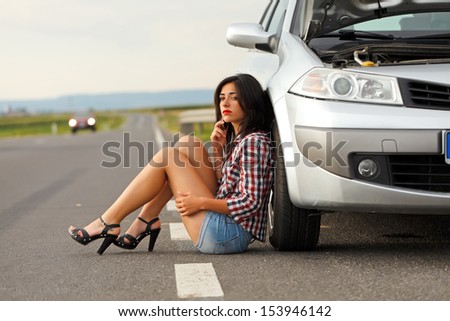 Sad woman sitting on the road near her broken car while talking on the phone