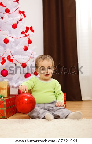 Happy little boy sitting in front of Christmas tree, holding big red ornament