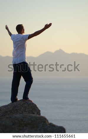 Man stands on a rock by the sea with his arms raised to the sky