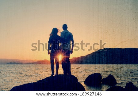 Young couple standing on the shore in the arms and looks at the setting sun by the sea. Filtered image: vintage, grunge and texture effects
