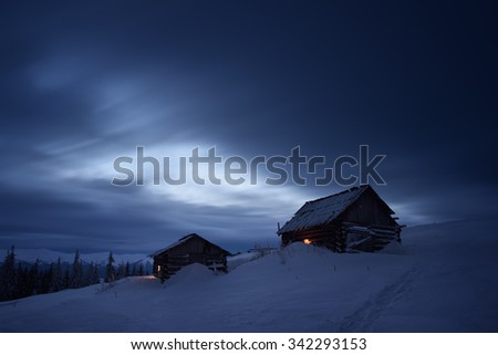 Night landscape in the mountains. Two wooden houses in the mountain settlement. Light in the windows. Path in the snow. Cloudy sky. Carpathians, Ukraine, Europe