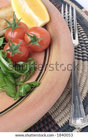 A glazed stoneware plate with Salad and fork on an African woven table mat. Some grilled seafood in the background