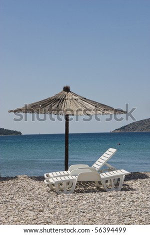 Big thatched beach umbrella and plastic chairs on rocky beach with open sea and islands in background